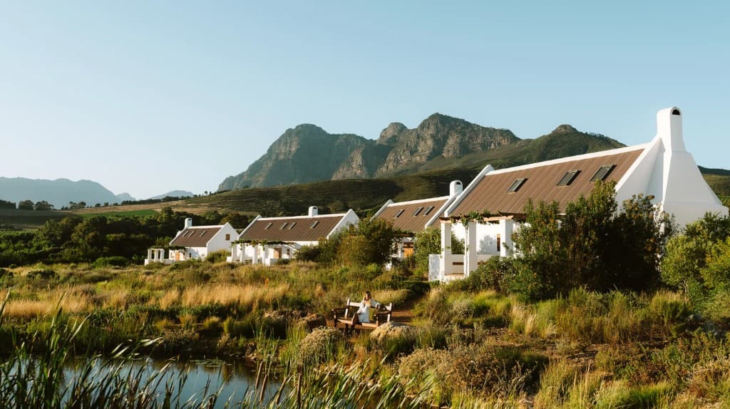 A guest sitting on a bench by the farm dam at Babylonstoren, looking toward the mountains and Cape Dutch cottages